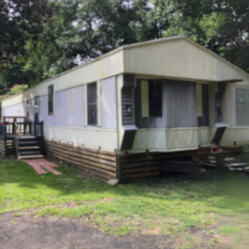 An older single-wide mobile home in need of repair, shaded by trees
